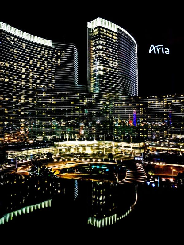 Aria večerní pohled na hotel Aria v Las Vegas, budova osvětlená světly, moderní architektura, skleněné fasády odrážející se ve vodní ploše před hotelem, jemné noční osvětlení a měkké odlesky na hladině vody