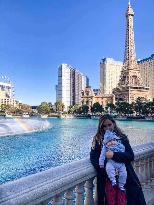 Baby žena drží miminko v náručí před fontánou, v pozadí je Eiffelova věž v Las Vegas, slunečný den, jemné odlesky vody ve fontáně, atmosféra turistického místa, turisté v dálce