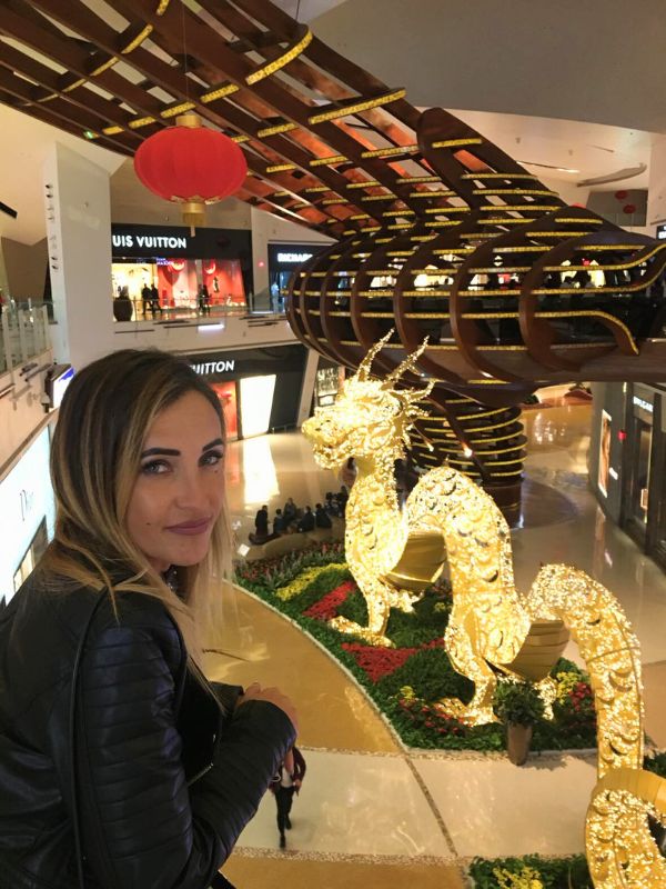 Dragon žena uvnitř nákupního centra v Las Vegas, nad ní velký osvětlený drak visící ze stropu, barevná světla a moderní architektura centra, luxusní obchody v pozadí, turistická a živá atmosféra, pohled zaměřený na dekoraci i návštěvnici