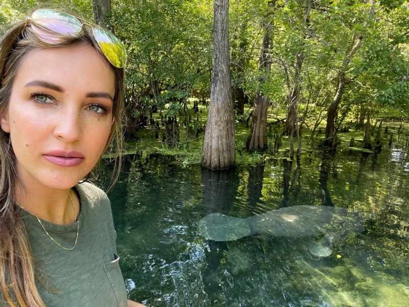 Osoba si dělá selfie u průzračného pramene obklopeného stromy. Ve vodě je vidět kapustňák, což vytváří klidnou a poklidnou atmosféru.