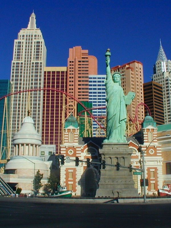 New York hotel New York-New York v Las Vegas, panoramatický pohled na mrakodrapy připomínající newyorskou architekturu, horská dráha vinoucí se kolem budov, socha Svobody v popředí, slunečný den, turisté a rušná městská atmosféra, živé barvy a detaily tematického resortu