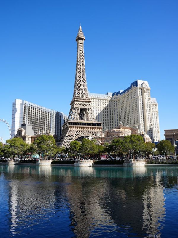 Paris Paris Eiffelova věž v Las Vegas mezi hotely, v popředí vodní plocha s jasným odrazem věže, večerní nebo noční osvětlení, světla hotelů se zrcadlí ve vodě, turisté a ruch města v pozadí.