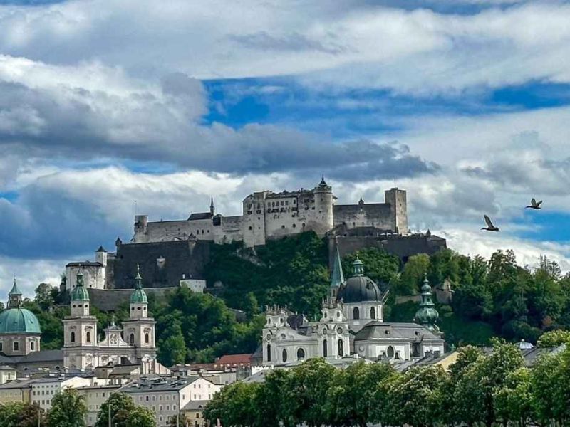 Středověký hrad stojí na vrcholu zeleného kopce a shlíží na historické město s honosnými kopulemi. Pod dramatickou oblohou plnou mraků létají ptáci.