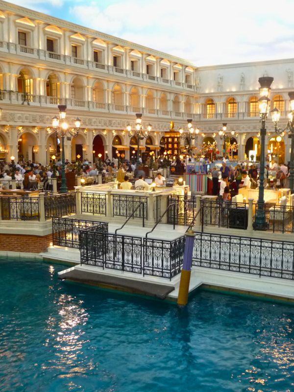Shopping uvnitř hotelu Venetian v Las Vegas, interiér s restauracemi a obchody, strop malovaný jako obloha, kanál s klidnou vodou, gondoly a lehce viditelní turisté, romantická atmosféra připomínající Benátky, elegantní dekorace a jemné osvětlení