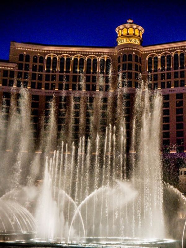 The Bellagio hotel Bellagio v Las Vegas v noci, budova osvětlená teplým světlem, před hotelem fontána stříkající vysoké vodní trysky, odlesky světel ve vodě, noční městská atmosféra s jemnými neonovými světly v pozadí