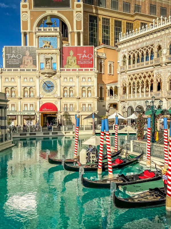 The Venetian Hotel hotel v Las Vegas inspirovaný Benátkami, italská architektura s pastelovými fasádami a oblouky, před hotelem vodní kanál s typickými gondolami, jemné odlesky vody, slunečný den, turisté po boku kanálu, atmosféra romantických Benátek přenesena do Las Vegas