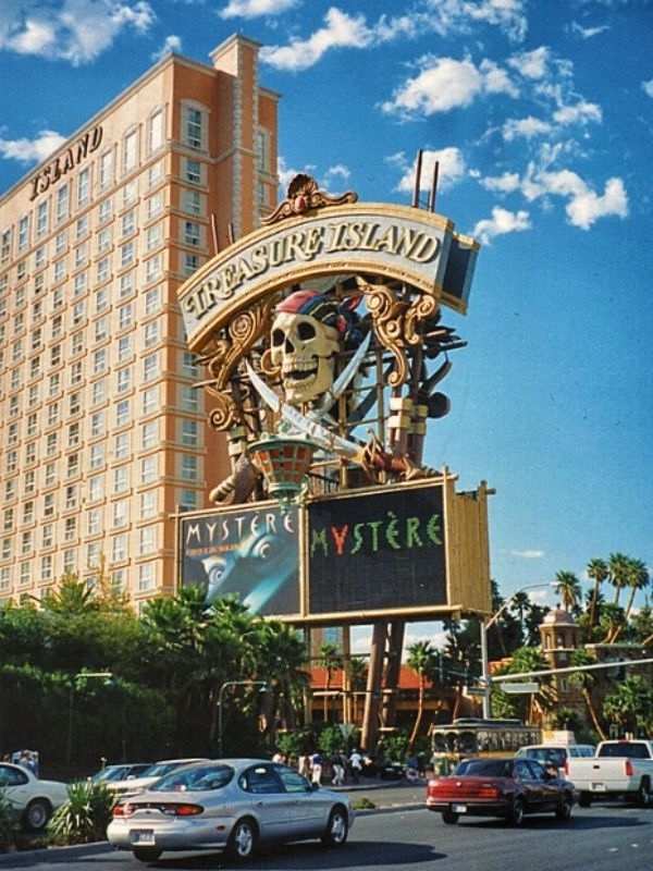 Treasure Island Treasure Island Hotel v Las Vegas, na fotce výrazné logo hotelu s pirátskou lebkou, budova hotelu v pozadí, slunečný den, turisté a ruch před hotelem, typická tematická dekorace připomínající pirátské dobrodružství