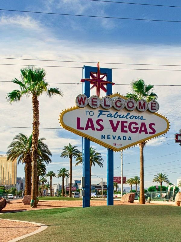 Welcome Sign velký uvítací nápis „Welcome to Fabulous Las Vegas Nevada“, obklopený palmami po stranách, jasně osvětlený večerními světly, nápis zářící proti tmavé obloze, pohled z mírného úhlu zepředu