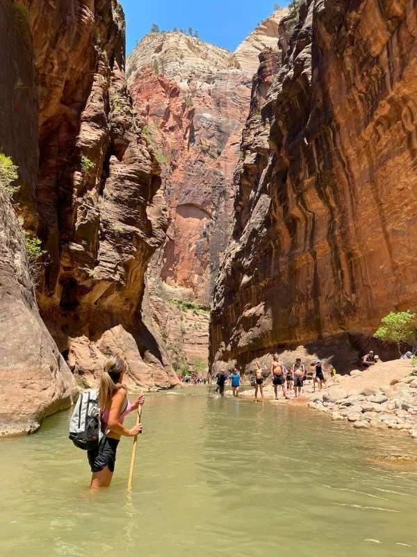 Turista s batohem a trekovou holí brodí úzkou řeku mezi impozantními červenými skalními útesy pod jasně modrou oblohou. Vpředu jsou vidět další turisté. Túra, nazývaná **The Narrows**, se nachází v **Utahu**.