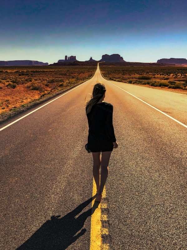 Dívka v černých šatech jde po dlouhé rovné silnici, v pozadí Monument Valley v Utahu.