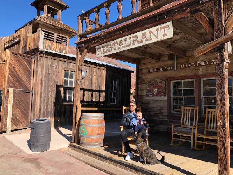 Muž se psem a dítě sedí před starou dřevěnou restaurací v Calico Ghost Town.
