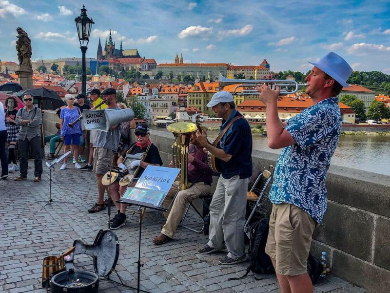 Pouliční kapela vystupuje na dlážděném mostě s malebným panoramatem města v pozadí. Hudebníci hrají na trumpetu a kytaru, zatímco kolemjdoucí přihlížejí.