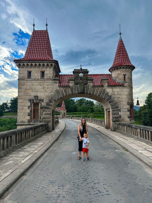 Žena a dítě stojí a usmívají se na kamenném mostě pod středověkým obloukovým mostem s věžemi se červenými střechami. Obloha je zamračená, což vytváří dramatické pozadí.