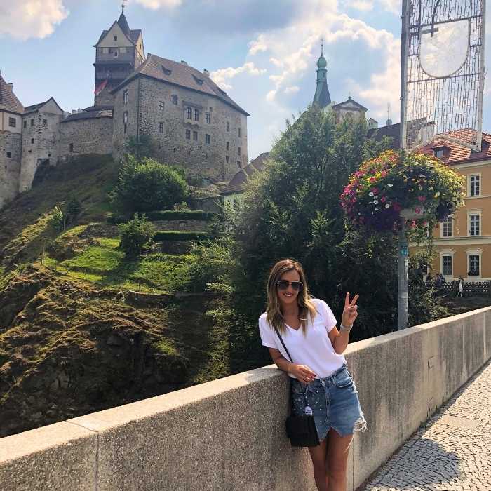 Žena v bílém tričku a sukni pózuje před hradem Loket.