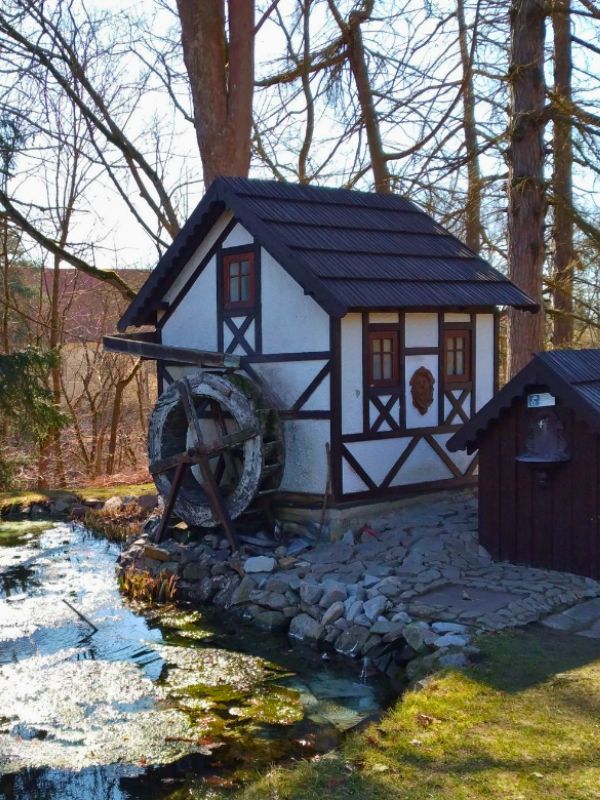 Malebný hrázděný vodní mlýn s oprýskaným kolem stojí u klidného potoka, obklopený stromy bez listí. Celá scéna vyvolává rustikální a poklidnou atmosféru.