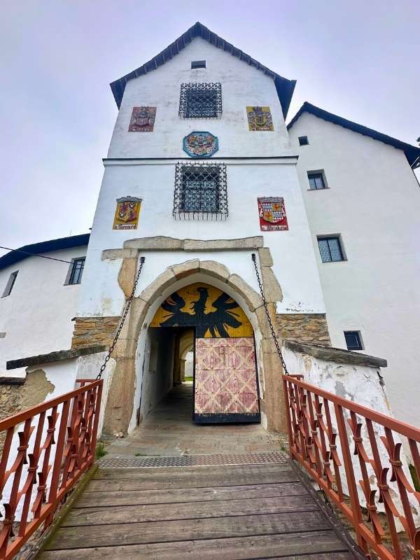 Dřevěný most vedoucí k historické bílé budově s klenutým vchodem. Fasádu zdobí erby, které dodávají místu pocit tradice a velkoleposti.