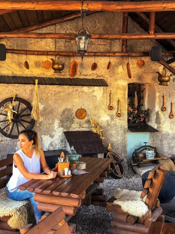 Žena v rustikálním prostředí sedí u dřevěného stolu pod dřevěnou pergolou. Výzdobu doplňují zavěšené měděné hrnce, kolo a sušená kukuřice. Atmosféra je útulná a uvolněná.