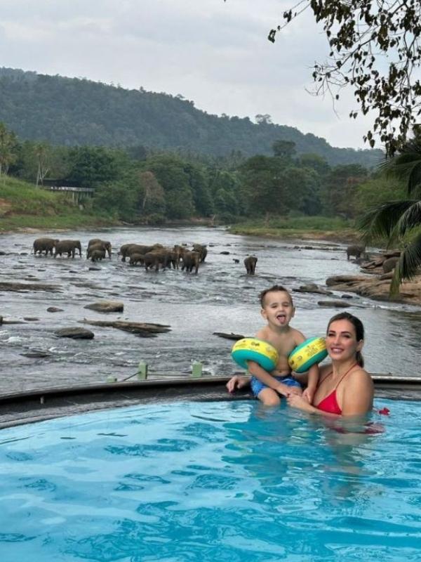 žena a malé dítě se usmívají do kamery, za nimi se koupající stádo slonů v řece