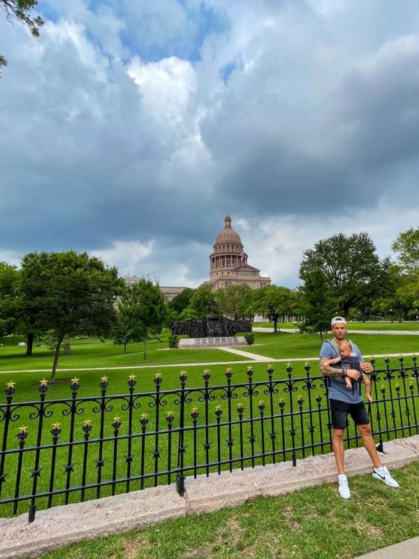 Hnědá budova v zeleném parku a otec, který drží malé dítě v nosítku, zamračená obloha.