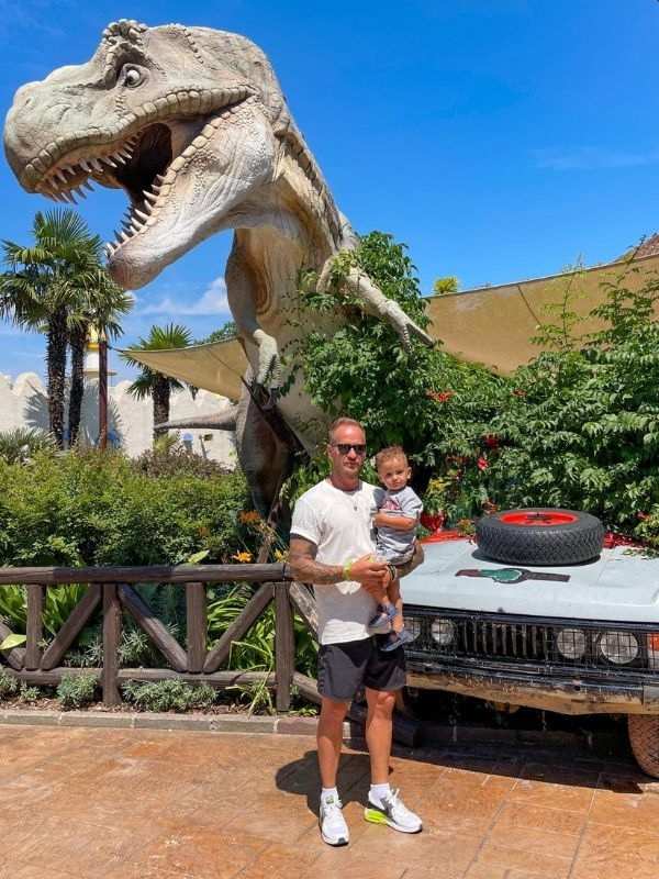 **Muž a malý chlapec pózují před autem, za nimi stojí velký dinosaur.**