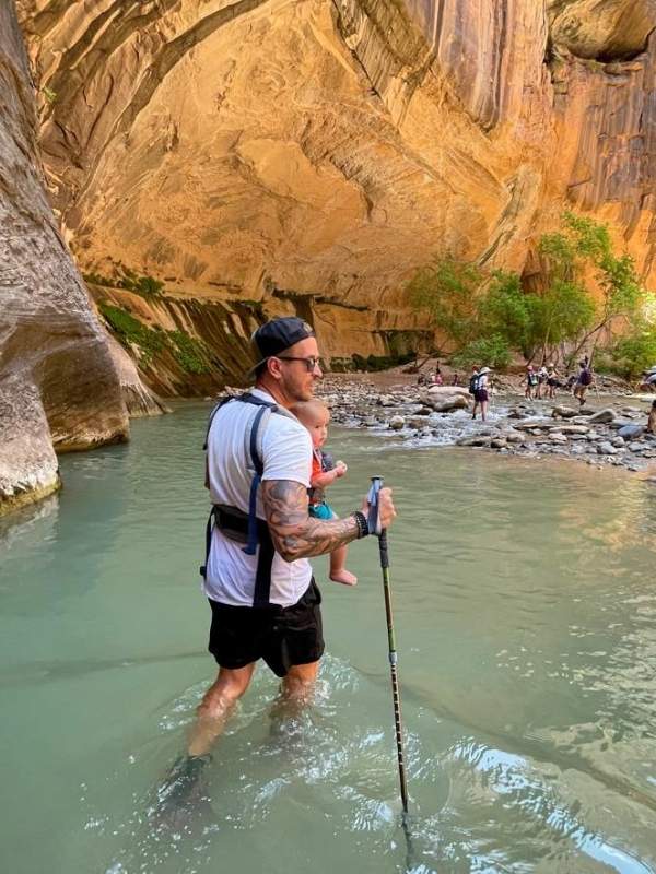 Hiking Narrows with Baby muž s batoletem v nosítku prochází vodou v kaňonu The Narrows v národním parku Zion, opírá se o hůlku, kolem vysoké skály