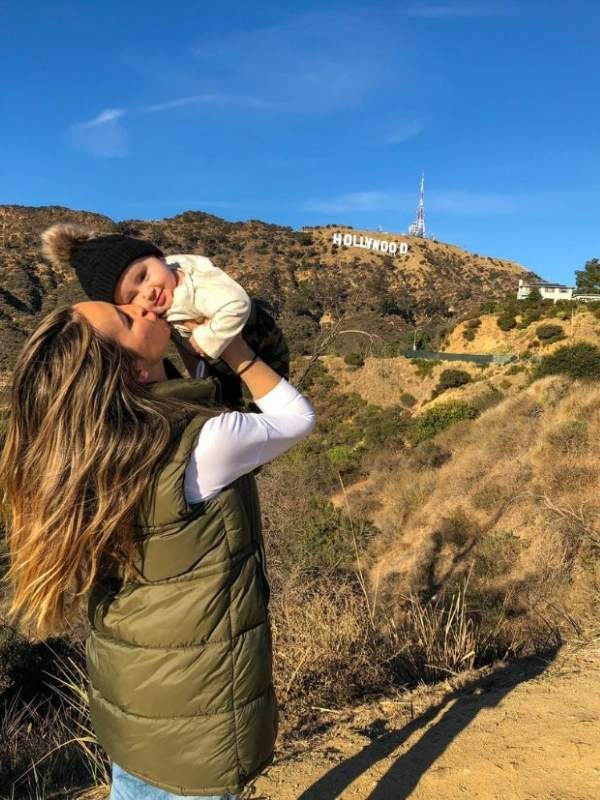 žena v zelené vestě dává pusu malému chlapci, v dálce nápis Hollywood na kopci