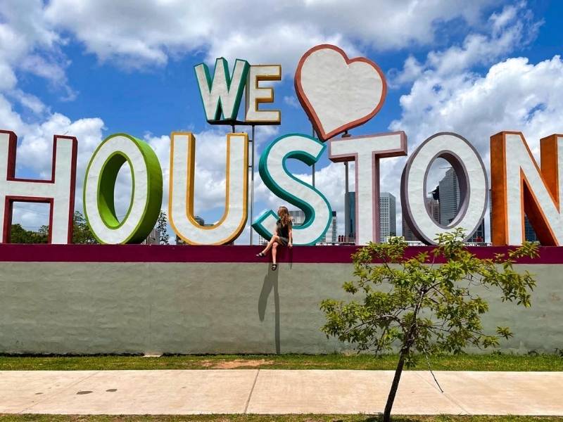 Žena sedí na zdi pod velkým nápisem We ♥ Houston.