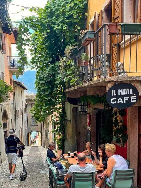 **Úzká italská ulička v Malcesine, pár lidí sedí u kavárny In Punto Cafe.**