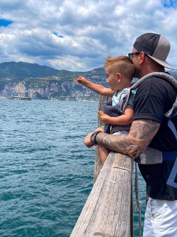 **Otec drží malého chlapce sedícího na zábradlí, oba se dívají do dálky na jezero a chlapec ukazuje.**