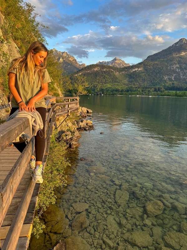 Žena sedí na zábradlí a dívá se do průzračné vody, v pozadí hory