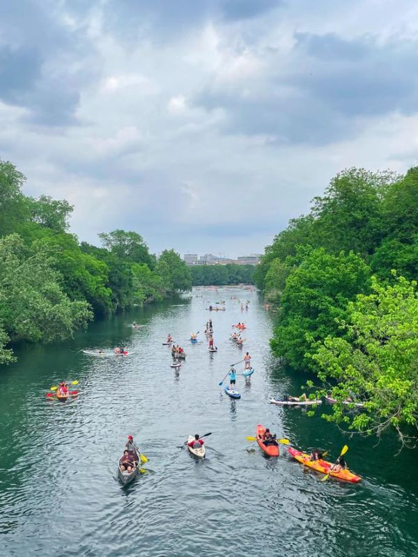 Řeka v Austinu plná kajakářů, kolem zelené stromy a zamračená obloha.