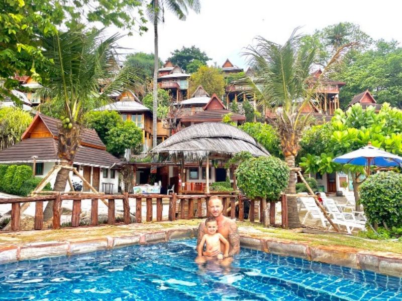 Otec se synem pózuje v bazénu, za nimi je resort rozprostřený do kopce