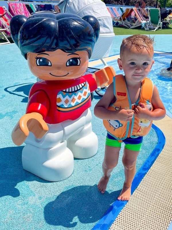 **Figurka z Lega a malý chlapec ve světlé vestě stojí v modré vodě bazénu.**