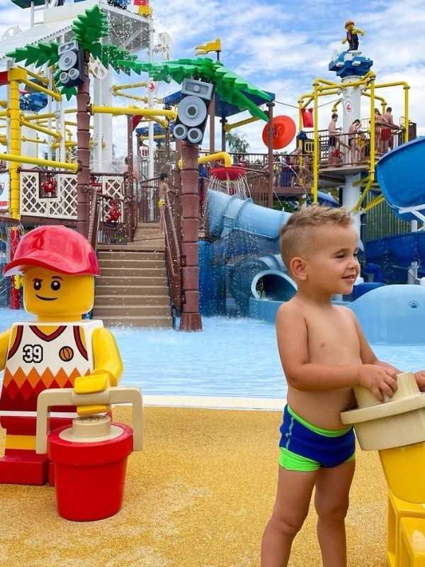 **Tobogány, prolézačky a bazén v Legolandu, v popředí žlutá figurka a smějící se chlapec.**