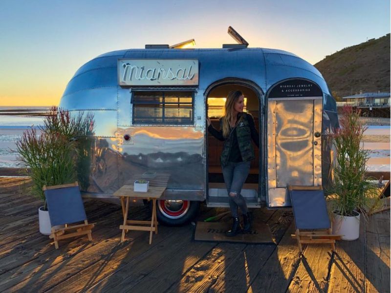 stříbrný karavan na Malibu molu, žena stojí ve dveřích, kolem jsou sedáky a květiny, s odrazem západu slunce