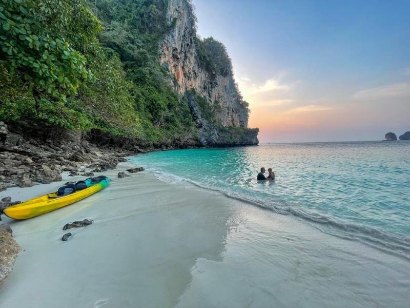 Dva lidé se koupou v průzračně modré vodě, na prázdné pláži stojí kajak a začíná západ slunce