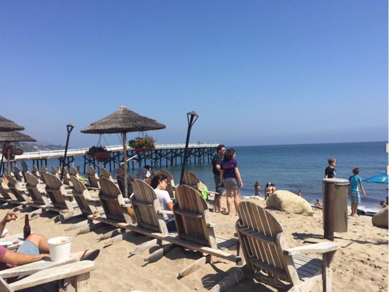 dřevěné židle v písku natočené k oceánu, tiki slunečník a molo v pozadí