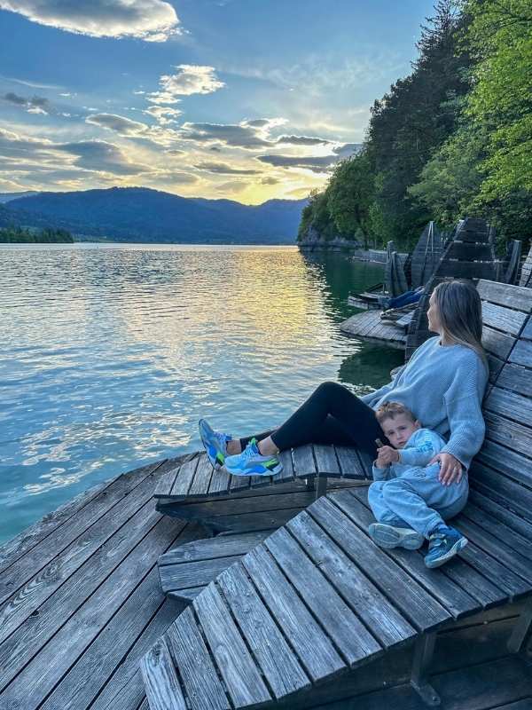 Žena a dítě sedí na dřevěných lavičkách a sledují západ slunce nad jezerem