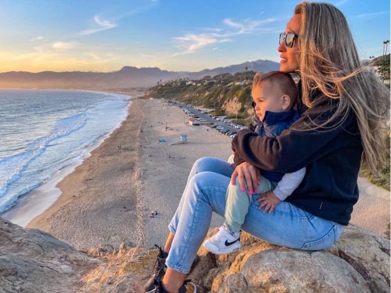 žena sedí se synem na klíně na kameni vysoko nad pláží s oceánem a sledují západ slunce v Kalifornii