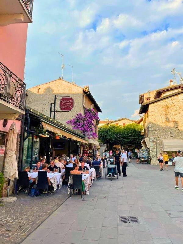 **Italská promenáda s kamennými domy, venkovní restaurací a lidmi u stolů.**