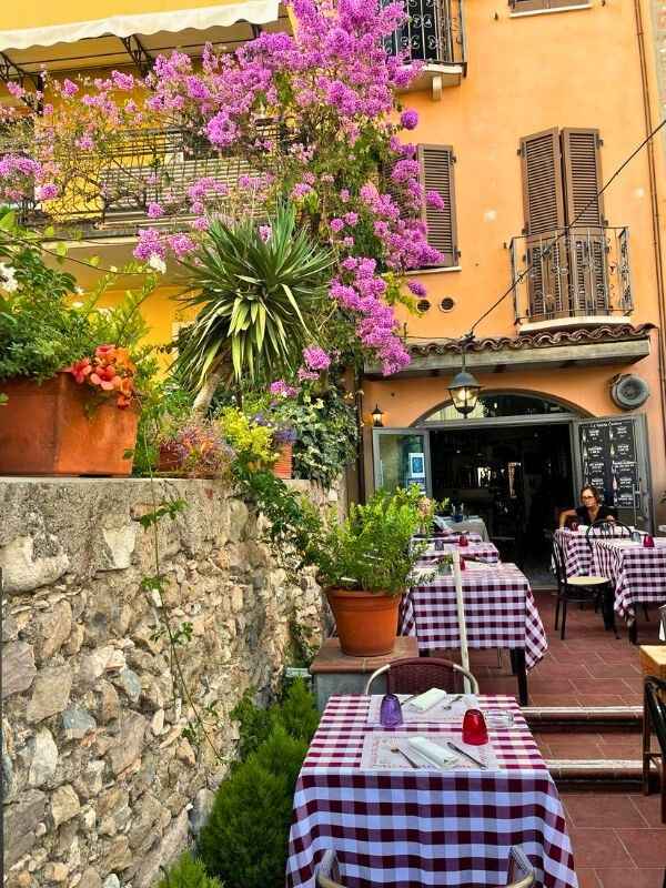 **Italská restaurace se žlutými zdmi, kostkovanými ubrusy a fialovými květy kolem.**