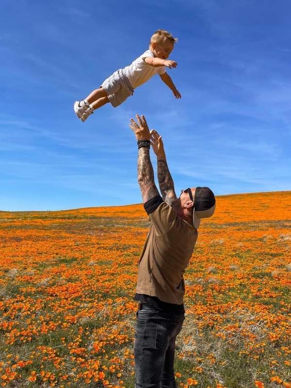 Otec vyhazuje syna do vzduchu uprostřed pole vlčích máků, modrá obloha v pozadí