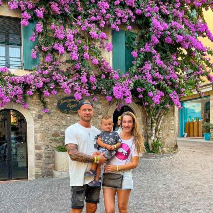 **Rodina se usmívá a pózuje na kameru před kamenným baráčkem s modrými okenicemi, obrostlým fialovými květy.**