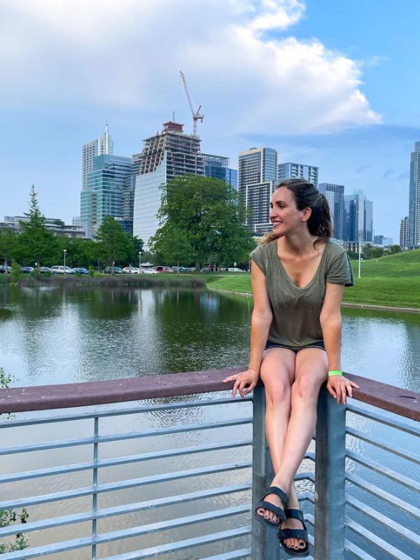 Žena sedí na zábradlí v parku, kolem voda a v pozadí obrysy mrakodrapů Austinu.