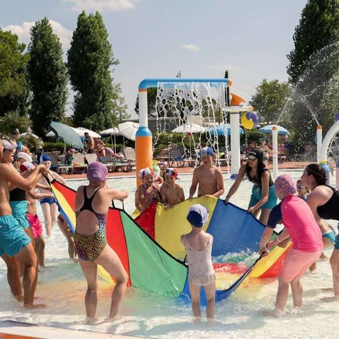 **Děti a dospělí drží velkou barevnou plachtu v bazénu, voda stříká a všichni si hrají.**