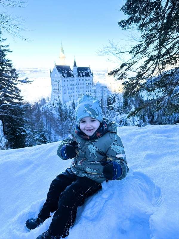 Chlapec se usmívá a pózuje ve sněhu, v pozadí zámek Neuschwanstein.