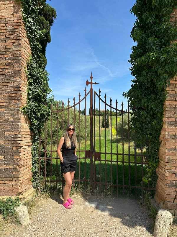 Žena stojí před železnou branou v Pienze pod modrou oblohou.