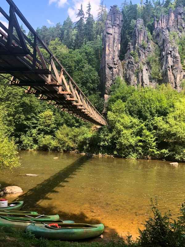 Dřevěný most přes řeku s kanoí v popředí a skalami a zelení v pozadí.