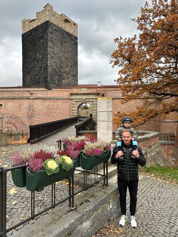 Vstup do hradu v Chebu s velkou černou věží, květinami a mužem nesoucím chlapce na ramenou.