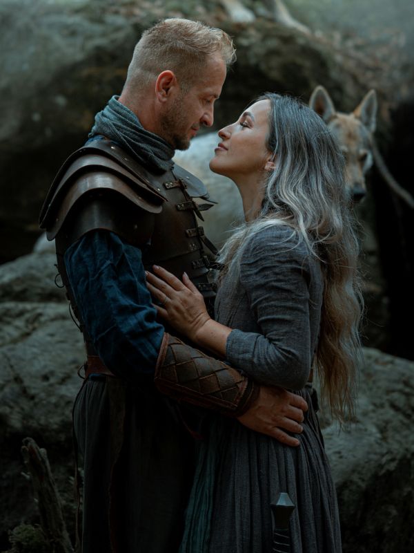 Dvojice ve starověkém oblečení se láskyplně drží, v pozadí je vlk